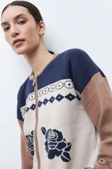 Chaqueta de tejido punto con cierre frontal, cuello redondo y manga larga. Presenta un diseño jacquard en tres colores: azul marino, beige y marrón camel. El cuerpo principal es beige con motivos de rosas azules y patrones geométricos en azul marino. Los hombros y mangas son de color marrón camel con patrones geométricos en azul marino. El cuello y el elástico inferior son de color azul marino.