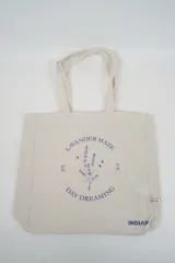 Bolso tote de tela con estampado de rayas verticales rosadas y blancas, laterales y asas en color beige. Presenta la palabra "BONJOUR" en verde en el centro y "INDIAN" en la parte inferior.