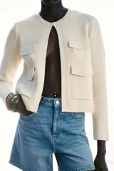 Chaqueta color crema de cuello redondo y manga larga, con bolsillos de parche con solapa en el frente y cierre frontal con corchete oculto.