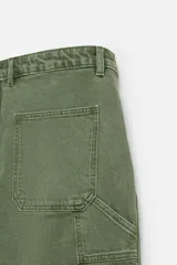 Bermuda color verde militar, de corte relaxed fit, con bolsillos frontales y traseros tipo plaqué, detalle de tira y bolsillo multifunción en la pierna, y bajo irregular.