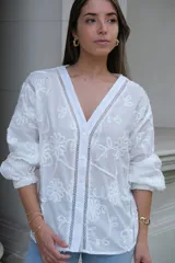 Camisa blanca con bordado de flores, cuello en V, cierre frontal con botones, mangas largas con puños elásticos y detalles de encaje.