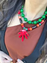 Collar corto con cuentas de coral, cerámica y acrílico en tonos naranja, rojo, marrón y verde. Dije de hoja pintado a mano en color rojo. Cierre con cadena de acero quirúrgico y ganchito para regular el largo.