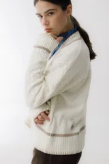 Sweater beige con cuello en V y mangas largas con diseño trenzado. Tiene dos bolsillos frontales con bordados de tréboles rojos y ribetes rojos en cuello, puños y ruedo.