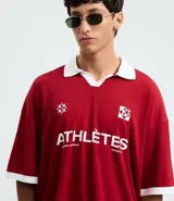 Remera de manga corta estilo polo, color rojo, con cuello y puños en contraste blanco. Presenta un corte oversized con hombros caídos y la palabra "ATHLÈTES" estampada en blanco en el frente. Incluye pequeños gráficos blancos a cada lado del pecho.