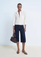 Camisa blanca de popelín de algodón, manga tres cuartos, corte crop y diseño recto. Presenta cuello camisero, bolsillo en el pecho con detalle de strass y cierre frontal con botones.