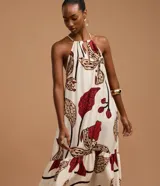 Vestido midi de corte evasé, con escote halter y abertura frontal en el busto, anudado en la parte superior. Presenta un estampado grande de flores y hojas en tonos rojo oscuro, beige y negro sobre fondo crudo. El diseño incluye un volante fruncido en el ruedo inferior.
