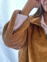 Buzo de pana fina color verde oliva, con diseño de corte holgado y mangas largas. Presenta una textura de cordón vertical característica de la pana y un pequeño parche decorativo en la parte superior de la espalda.