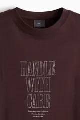 Remera color bordó de corte holgado con cuello redondo y mangas cortas. Estampado frontal con la frase "Handle with care".