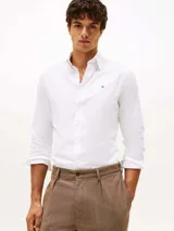 Camisa blanca de corte regular, confeccionada en tejido Oxford de algodón con un toque de elasticidad. Tiene cuello abotonado, mangas largas con puños abotonados y logo de Tommy Hilfiger bordado en el pecho.