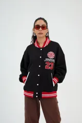 Campera varsity negra con detalles en rojo y blanco, cierre con botones a presión, cuello, puños y cintura acanalados con franjas a contraste, bolsillos ribeteados y aplique bordado en el pecho con el texto "Kingpand Letsurestyce 23".