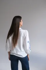 Camisa blanca de manga larga con elástico en puños, cuello mao con botones en la parte delantera y bordados florales azules en el frente y las mangas.