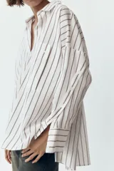 Camisa blanca con rayas verticales grises, de corte holgado, cuello solapa y mangas largas con puños abotonados. Tiene un bolsillo de parche en el pecho, pinzas en la espalda y bajo asimétrico con aberturas laterales.
