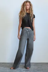 Pantalón de jean celeste de tiro alto, corte wide leg y largo hasta el tobillo. Presenta un diseño asimétrico en la cintura con cierre frontal cruzado con cremallera y botones metálicos.