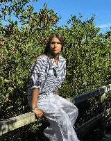 Camisa de manga corta con cuello y botones frontales, confeccionada en tela a rayas verticales blancas y azul marino. Presenta un calce holgado y volados en los puños.