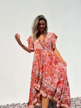 Vestido largo de corte evasé con estampado floral en tonos rosados y anaranjados. Tiene escote en V cruzado, mangas cortas con volantes y falda con volantes asimétricos.