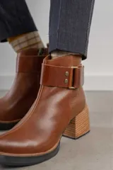 Bota corta de cuero marrón con puntera redondeada y taco cuadrado. Presenta una correa con hebilla alrededor del tobillo y cierre lateral.