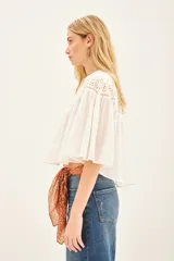 Camisa blanca de algodón con cuello redondo, corte amplio y mangas cortas abullonadas. Presenta detalles de calado en la parte superior delantera y trasera. Cierre frontal con botones.