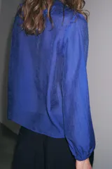 Camisa azul con escote en pico, cierre frontal con botones, mangas largas abullonadas con elástico en los puños y detalle de volantes en el frente.