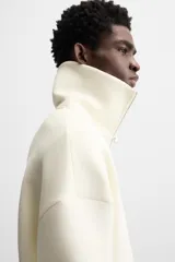 Sudadera color crudo de tejido técnico con cuello alto y cierre de cremallera. Tiene manga larga, corte relaxed fit, bolsillo tipo canguro y rib en puños y bajo con rayas negras.