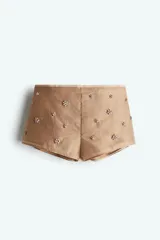 Short mini color beige en tejido liviano con detalles florales elaborados con strass. Modelo de cintura clásica con ribete fino de grosgrain y cierre oculto con cierre de corchete a un lado. Forrado.