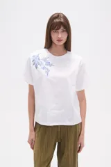 Remera blanca de manga corta y cuello redondo, con un bordado floral en tonos azules en la zona superior izquierda del pecho.