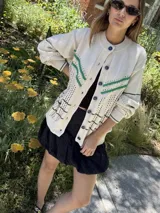 Saco de punto calado color beige claro, con cuello redondo y cierre frontal de botones azules. Presenta detalles decorativos en crochet verde y negro en el frente y puños.