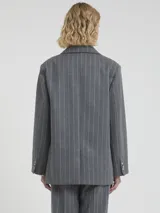 Blazer cruzado de lana gris con finas rayas blancas verticales (pinstripe). Presenta solapas anchas, doble botonadura con botones claros y un accesorio decorativo de cadena de perlas colgante prendido en la solapa izquierda.