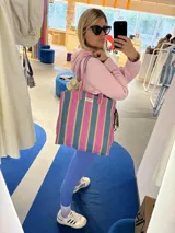 Bolso tipo tote bag con rayas verticales en tonos rosa, celeste y dorado. Tiene asas cortas rosas y etiqueta con la marca "AMÉN".