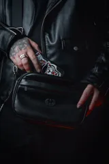 Bandolera negra de cuero sintético con correa ancha ajustable y desmontable. Cuenta con cierre superior, bolsillo frontal con tiracierre customizado, forro interno y cuatro bolsillos en total (dos exteriores y dos interiores).