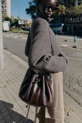 Bolso tipo saca de cuero sintético marrón oscuro, con fruncido superior ajustable mediante cordón. Posee asa de hombro con detalle de cadena metálica oscura y bolsillo interior con cierre.