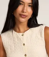 Blusa femenina de corte regular confeccionada en tejido de anarruga texturizado color beige. Presenta cuello redondo, diseño sin mangas y cierre frontal con botones.