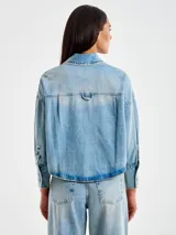 Chaqueta camisera de denim celeste, corta por delante y más larga por detrás, con cierre frontal con botones y bolsillo frontal aplicado en el pecho.