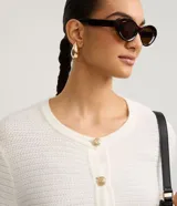 Cardigan de punto blanco, con cuello redondo, manga larga y cierre frontal de botones dorados. Posee dos bolsillos cuadrados en la parte delantera inferior.