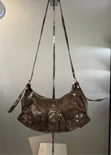 Cartera marrón con textura de cuero, forma de media luna y correa de hombro ajustable. Tiene cierre superior con cremallera y un bolsillo frontal con hebilla decorativa.