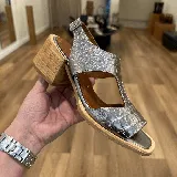 Sandalia plateada con textura tipo piel de cocodrilo, con talonera y tira ancha sobre el empeine con aberturas laterales. Tiene taco de madera y suela color suela.