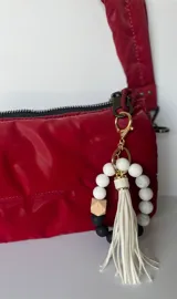 Llavero con pulsera de cuentas redondas de silicona en colores blanco, gris y negro, con una cuenta de madera hexagonal y aro metálico dorado. Incluye borla de flecos de cuero sintético color blanco.