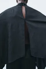Capa negra con cuello perkins, manga larga y bajo asimétrico. Detalle de aplicación de abalorios a tono y tejido arrugado. Cierre en espalda con abertura y botones.