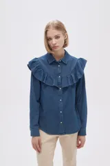 Camisa de jean azul con cuello clásico, cierre frontal con botones y mangas largas. Presenta volados en el canesú delantero y trasero.