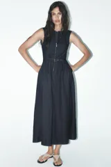 Vestido midi de denim azul oscuro, sin mangas, con cuello redondo, bolsillos laterales, cinturón con hebilla metálica y cierre frontal con cremallera.