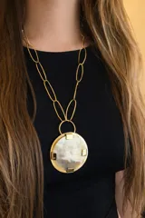 Collar con cadena de eslabones ovalados grandes en tono dorado. El dije es un medallón circular con borde dorado y centro de guampa (cuerno) en tonos beige y blanco, asegurado con cuatro piezas metálicas en forma de 'L' invertida.