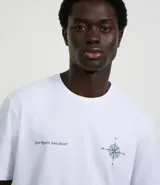 Remera blanca de algodón con corte clásico, cuello redondo y mangas cortas. Presenta estampado frontal con una brújula, un mapa y la inscripción "Sun Route Sailboat".