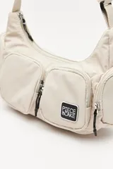Cartera tipo shoulder color beige, de diseño utilitario y formato curvo. Confeccionada en material liviano y resistente, cuenta con correa regulable para llevar al hombro o cruzada. Presenta múltiples bolsillos con cierre en el frente y laterales, y un parche con el logo "Piece & Cake" en el frente.