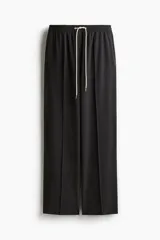 Pantalón negro de sarga suave con cintura elástica, cordón de ajuste en color contrastante y franjas laterales finas. Modelo de corte holgado con tiro alto, bolsillos laterales discretos y bolsillos insertados decorativos atrás. Piernas amplias con pliegues adelante y atrás.