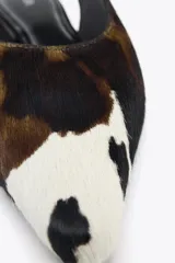 Zapato tipo bailarina destalonada en piel con pelo, con estampado animal print de vaca en blanco, marrón y negro. Tiene tira trasera con pieza elástica y acabado en punta. Suela plana de 1 cm de altura.