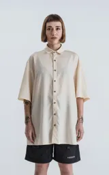 Camisa oversize color crema con cuello clásico, cierre frontal con botones marrones y mangas cortas.