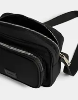 Bandolera negra de tela deportiva con bolsillo frontal con cierre, correa regulable, cierre superior y tag de goma.