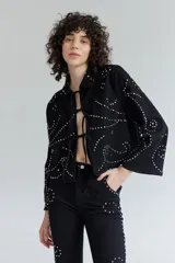 Chaqueta corta de denim negro con cuello clásico y cierre frontal con cordones. Decorada con tachas plateadas formando patrones ondulados.