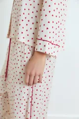 Pijama de dos piezas, compuesto por una camisa de manga larga y pantalón largo, ambos en tejido de viscosa color blanco con estampado integral de corazones rojos. La camisa tiene ribetes rojos en cuello, puños y abertura frontal. El pantalón tiene un fajín ancho color rojo liso.