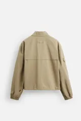 Cazadora color arena de corte holgado, con cuello solapa y manga larga con bolsillo multifunción. Tiene bolsillos frontales de plastrón, bolsillo interior y elástico en los puños y ruedo. El ruedo es ajustable con cordón. Cierre frontal con cremallera oculta por solapa con botones a presión.
