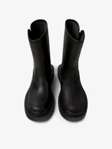 Bota de media caña color negro, con diseño de suela gruesa y estriada. Presenta una correa con hebilla en la parte superior de la caña y un logo de la marca "Camper" en una etiqueta.